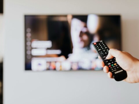 Im Hintergrund ist ein an der Wand befestigter Smart-TV zu sehen. Zentral im Vordergrund ist eine Hand zu sehen, die eine Fernbedienung hält und auf den Fernseher zeigt.